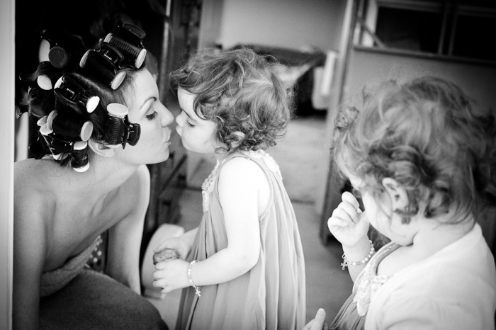 Documentary-style, black and white image of bride kissing her twin daughters during bridal preparations.